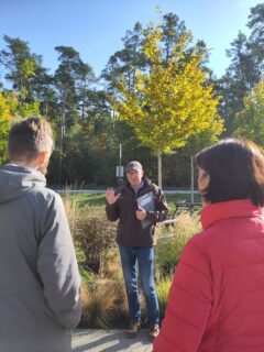 Der technische Leiter des Botanischen Gartens, Herr Heuvemann, mit interessierten Teilnehmern der Führung. Im Hintergrund eine Wiese mit Bäumen.